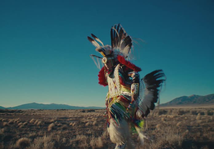 A person in colorful traditional regalia dances against a backdrop of open plains