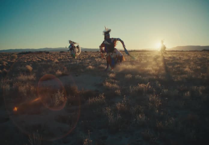 Three people wearing colorful feather costumes are dancing and spinning in a desert region