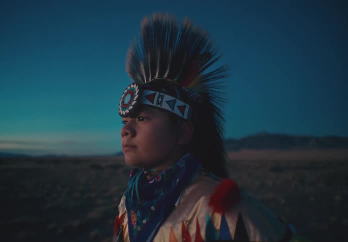 A young person standing in a desert region wearing an outfit with a feather cap