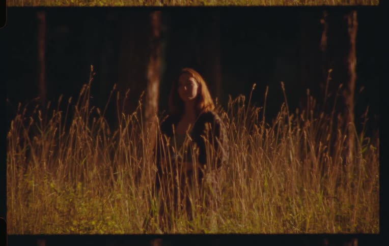 A woman is in a field with tall beige grass and she is pointing her camera forward and then taking it away from her face
