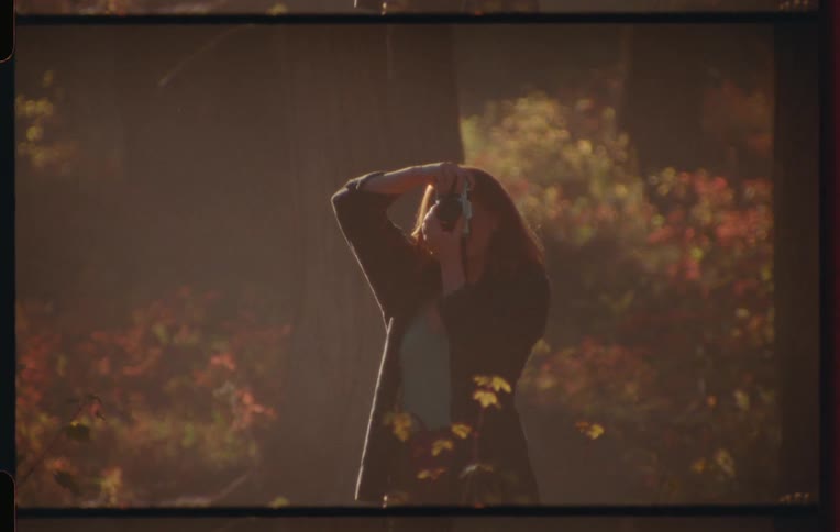 A woman with shoulder length brown hair holding a camera above her head taking a picture amidst an out of focus background of autumnal foliage