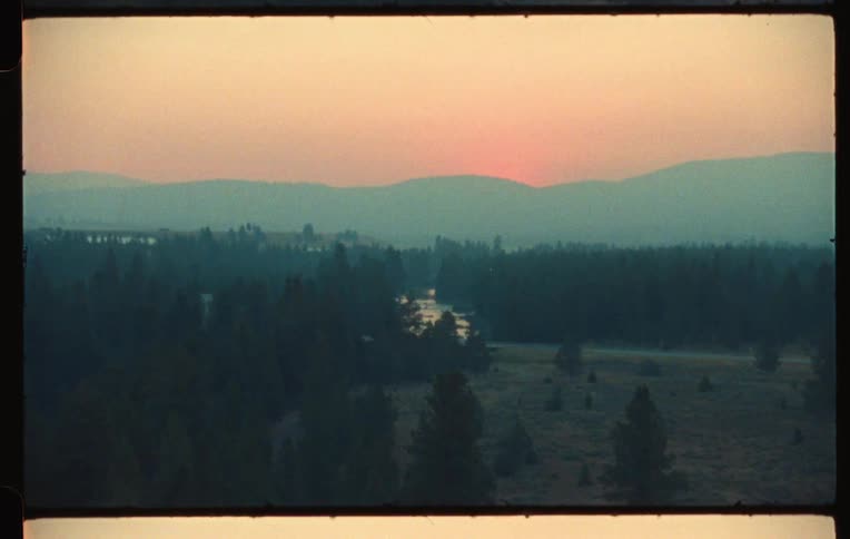 River winds through a conifer forest near foggy mountain under a red sky