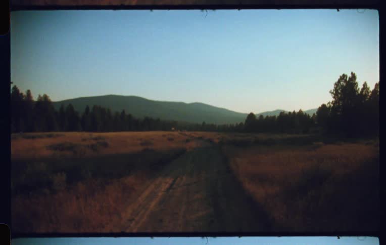A long dusty road winds through a field of dry grass and scrubby vegetation toward a line of mountains