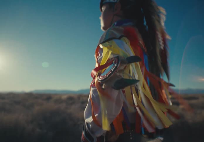 Man wearing a native american costume stands in a field on a windy day while looking towards the sun