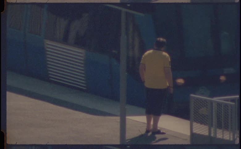 A person in a yellow shirt and dark shorts waits at a train platform as a blue train pulls up
