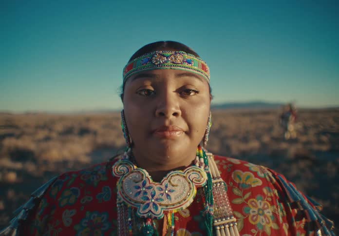 Woman wearing traditional native american attire stands in a field while another performer moves in the background during a traditional ceremony