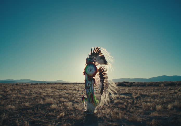 Man wearing native american attire turns while standing backlit in a field