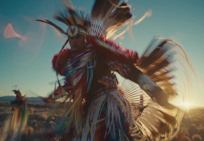A desert region with three people wearing clothes with various colorful feathers and they are doing a ritualistic dance