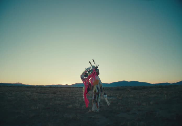 Person standing in a field wearing traditional native american clothing with the silhouette of mountains in the background