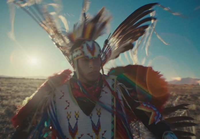 A person is dressed in traditional colorful native american regalia with feathers and intricate patterns dancing in an open field