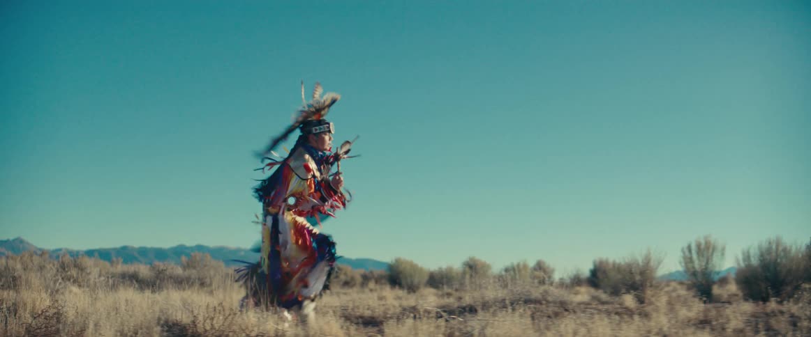A person is dressed in traditional colorful native american regalia with feathers and intricate patterns dancing in an open field