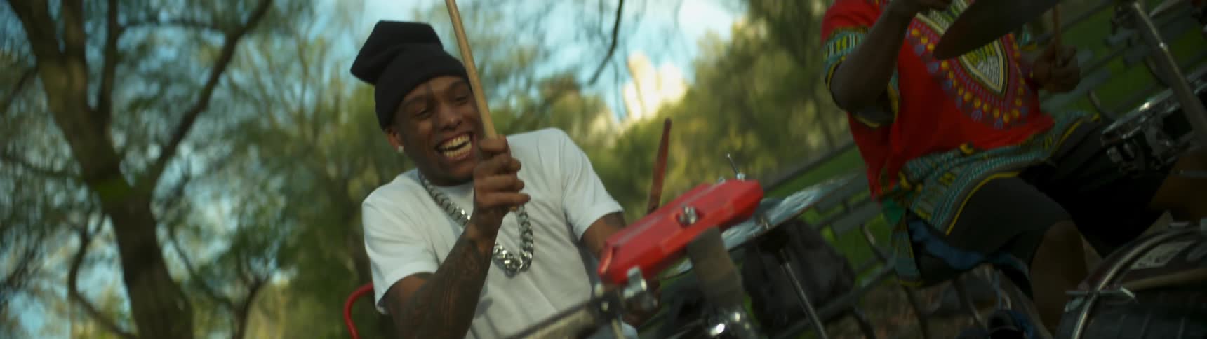 One guy is playing a drum with another guy sitting next to him in a park