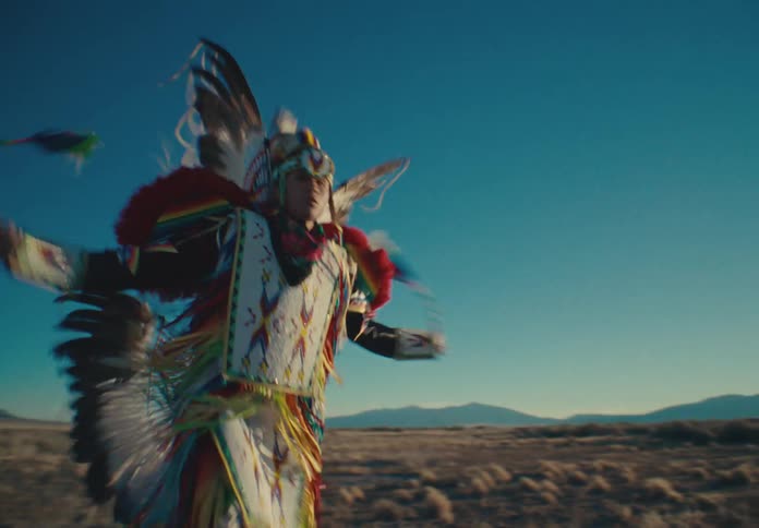 A person in colorful clothes and a feathered hat who is dancing by moving his hands forward and to the sides in a desert region