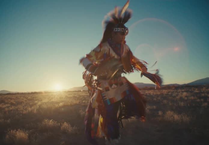 People in traditional native american attire dance during a ceremony on a plain lit by the sun shining from horizon