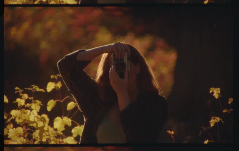 A woman with long reddish brown hair takes a photograph with a camera partially obscured by foliage in warm golden light