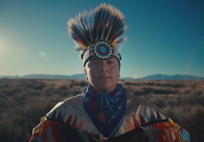 Girl in native american tribal attire stands in a field with mountains in the background