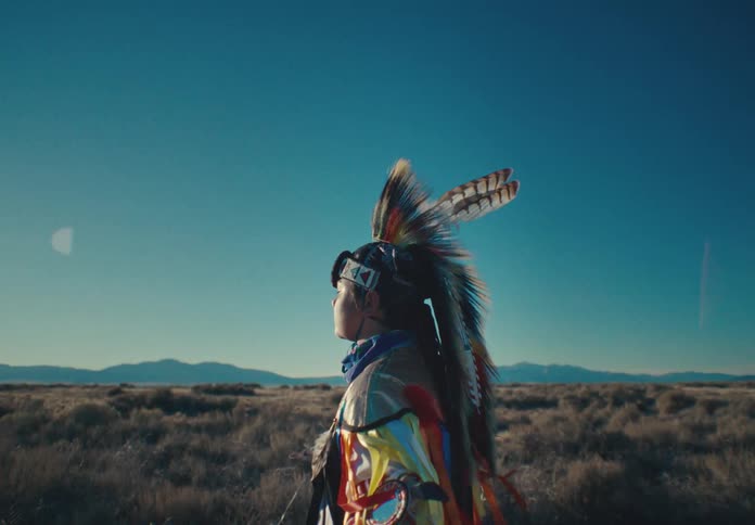 Person wearing traditional native american attire stands alone in a field on a windy day