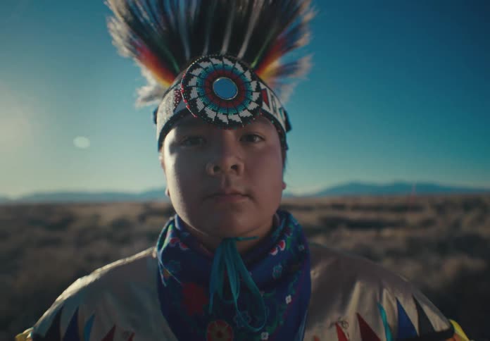 A young person wearing a traditional native american headdress and regalia looks directly at the camera against a desert landscape