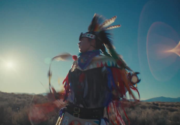 A person is dressed in traditional colorful native american regalia with feathers and intricate patterns dancing in an open field