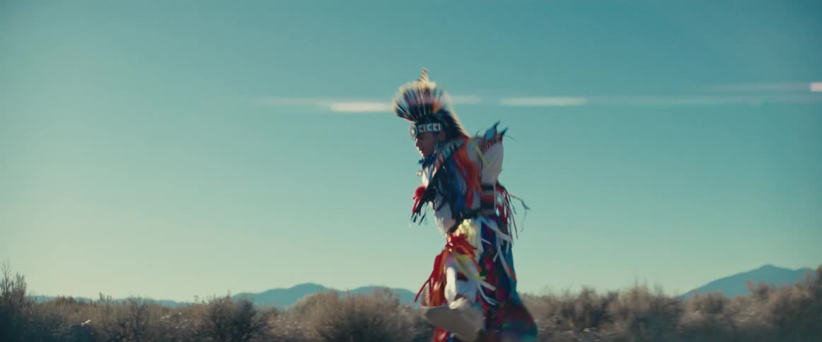A person in vibrant traditional native american regalia dances in a dry open landscape under a clear sky