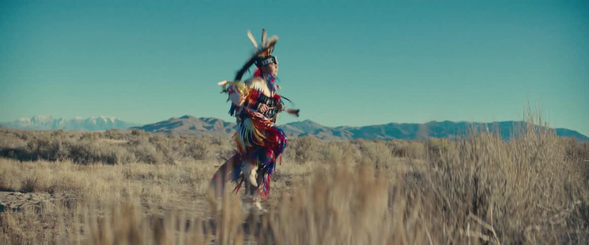 Person in native american costume dancing in a field with mountains in the background