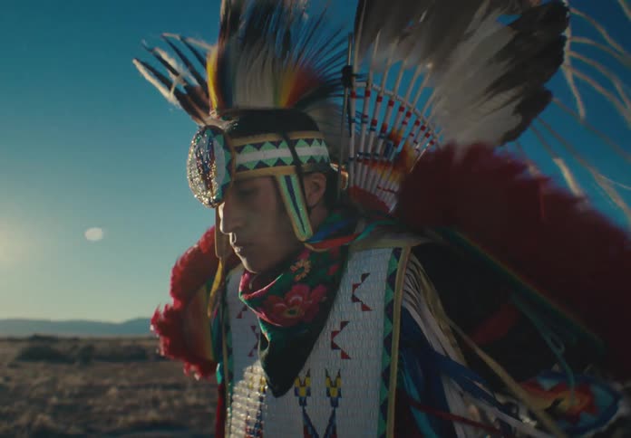 A person in traditional native american regalia stands in a dry open landscape bathed in the sunlight