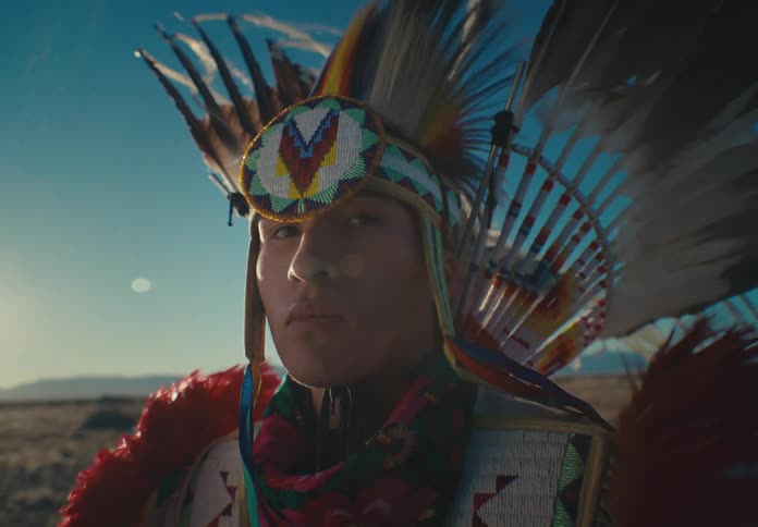 Portrait of a seriouslooking man wearing native american attire while standing in a desert setting with vegetation and mountains in the background