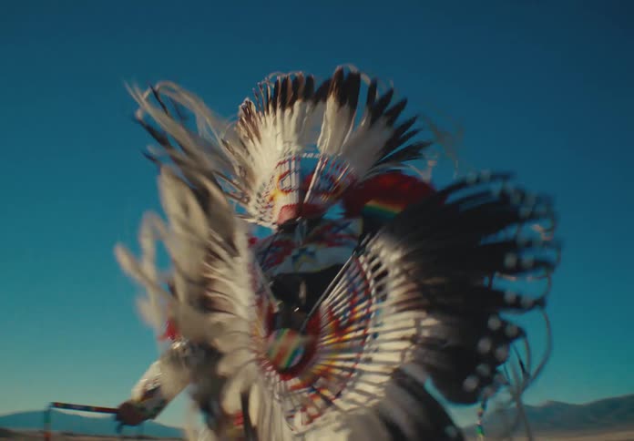 Native american man wearing traditional dress while dancing in a field with mountains in the background