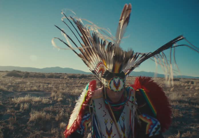 A person crouching in a desert area and wearing loose clothing made of colorful feathers is twirling two objects in his hands that have feathers on the ends