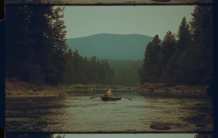 People ride in a rowboat floating on a river passing through a conifer forest with a fog obscured mountain in the background