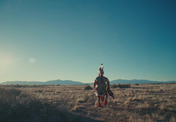 A person in colorful feathered clothing with a large hat is performing a ritualistic dance in a desert region