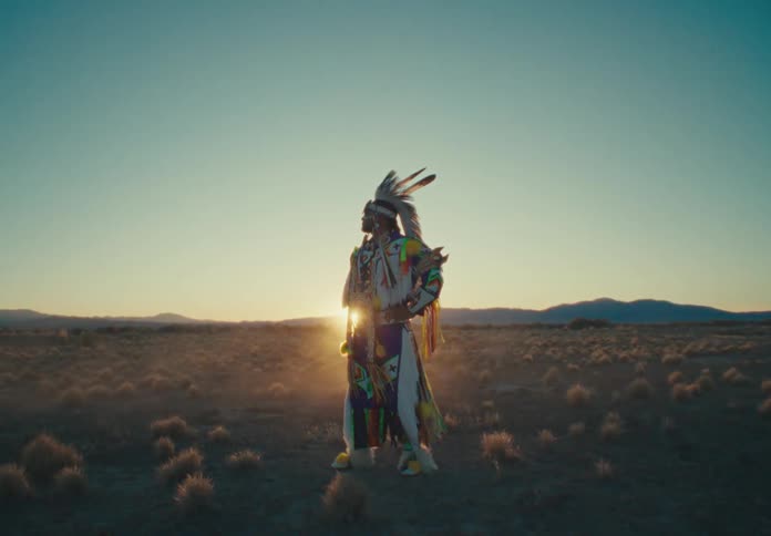 American inactive tribal man in traditional dress stands in natural setting with sunrise on the horizon