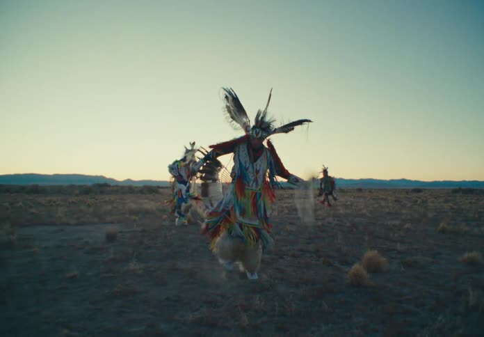 Several people in colorful native american regalia are dancing in a dry open field at sunrise or sunset