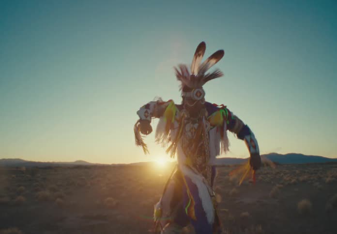 A person is dressed in traditional colorful native american regalia with feathers and intricate patterns dancing in an open field