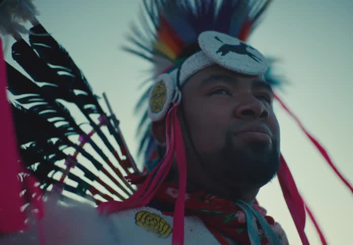 The face of a man who is staring straight ahead and he is wearing an outfit that is made up of feathers and he is wearing a big hat