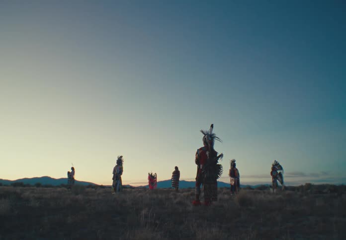 Seven people in large feathered clothing who are standing in a desert region and are immobile