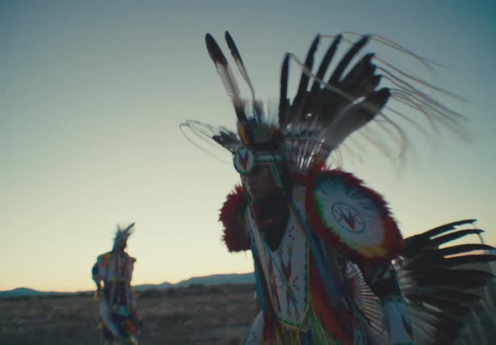 Two native american dancers in elaborate feathered headdresses and regalia as they move across a desert landscape at either dawn or dusk