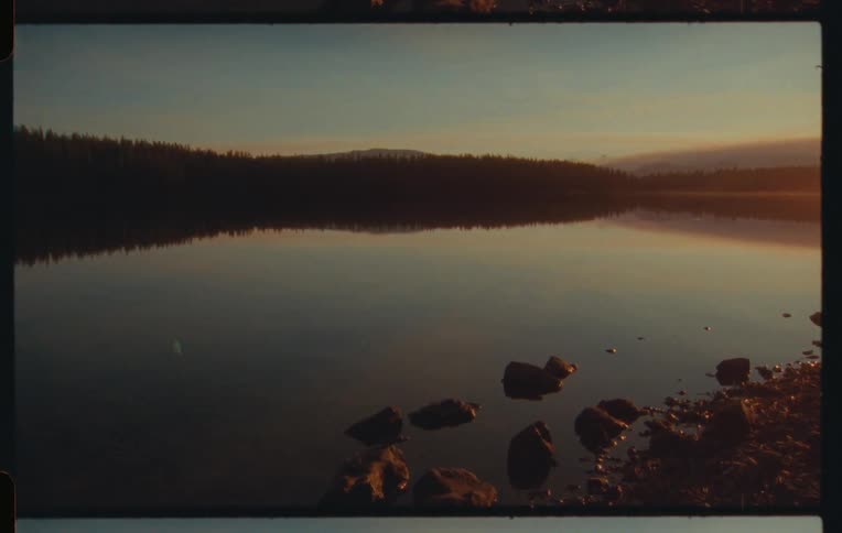 A calm lake at either dawn or dusk reflecting the muted colors of the sky and the dark silhouette of a tree line on the far shore