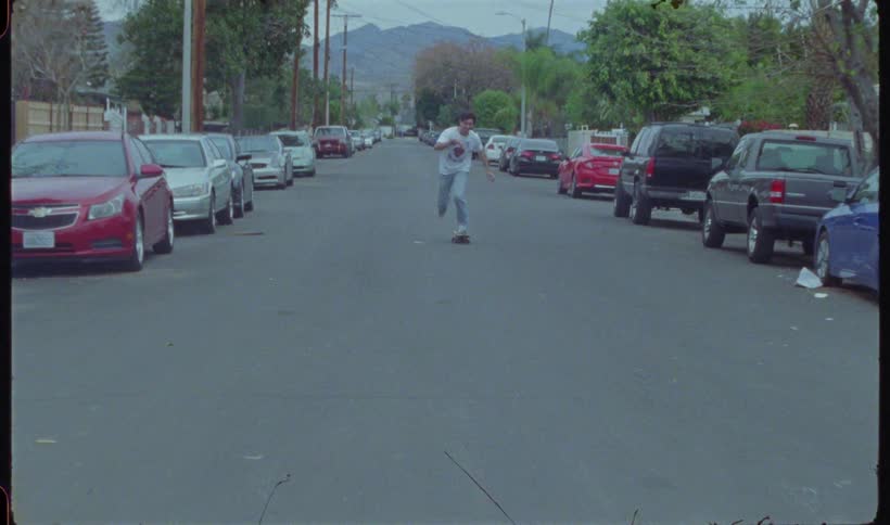 A young man is skateboarding on an asphalt street with cars parked on both sides