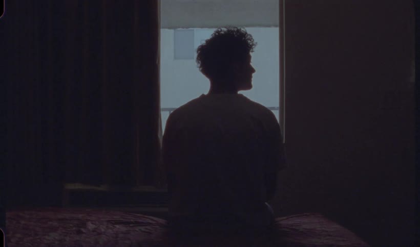 A young man sitting on a motel room bed with shoulders hunched lifts his head momentarily