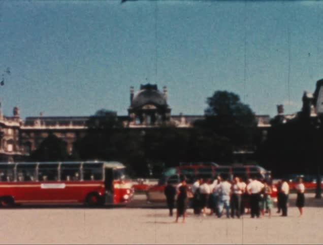 Group of people standing together talking in a public square with vintage red buses in the background