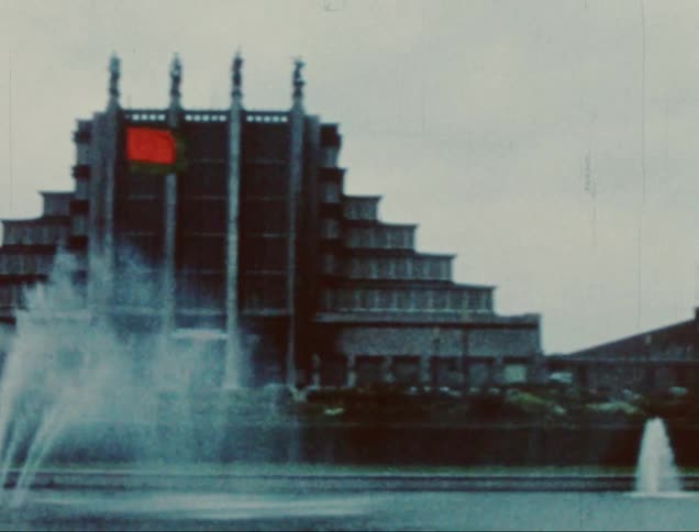 A young woman stands in front of a large building with multiple fountains in the foreground