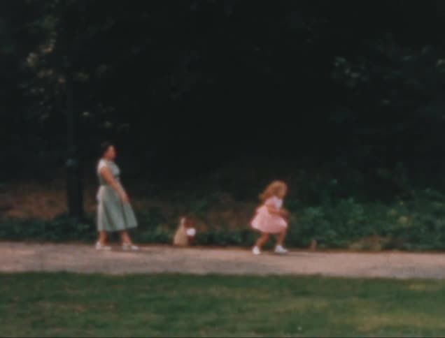 An old image of a little girl in a pink dress playing ball with an adult woman on a concrete path next to a grass field