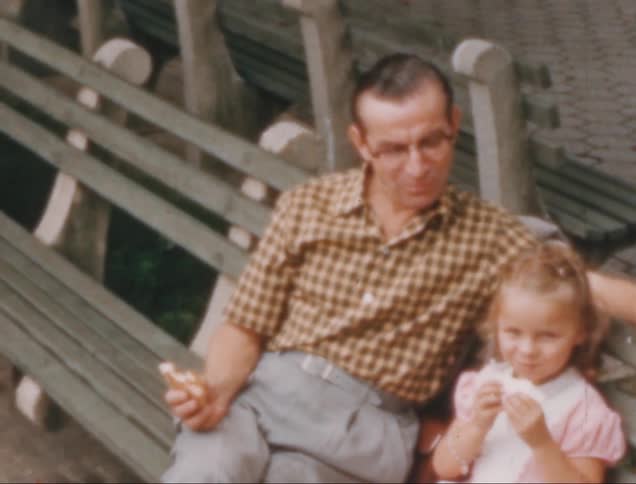 A father in a checkered shirt sitting on a bench next to a smiling daughter eating pastries in a white and pink dress outdoors in a park setting