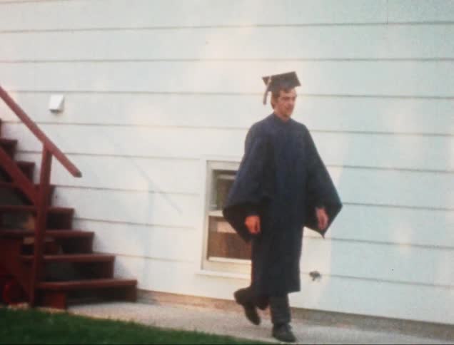 A young graduate in a cap and gown walks outdoors towards an elderly woman