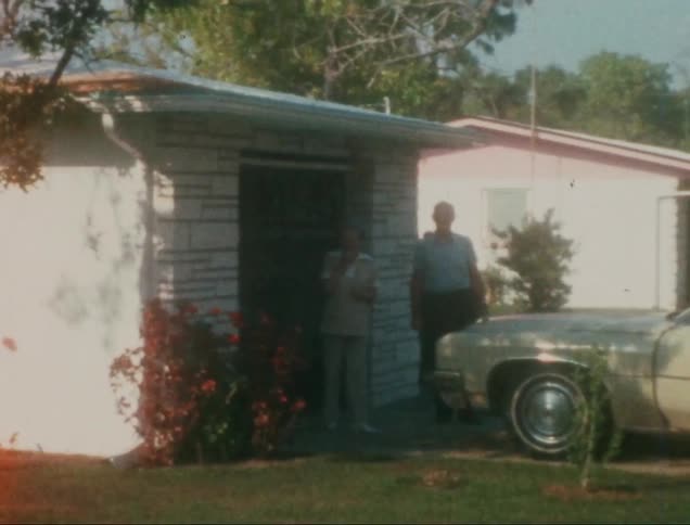 Two people are standing outside a small house with a white exterior next to a parked vintage car