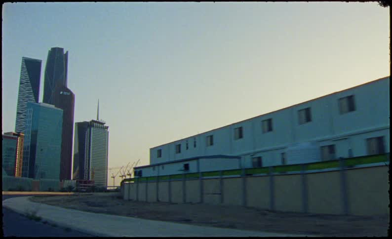A car drives down a road past a building to reveal the sun setting in the background