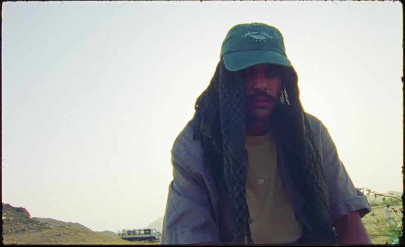 A man in a green cap with a piece of cloth underneath sits on an abandoned car in the countryside