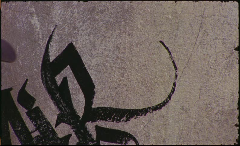 A hand with a watch on the wrist is holding a pen and painting a large arabic calligraphy on a concrete wall