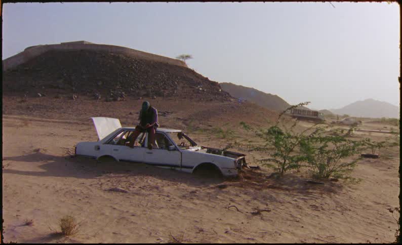 Man sitting rooftop on an abandoned half buried car in the desert on a windy day